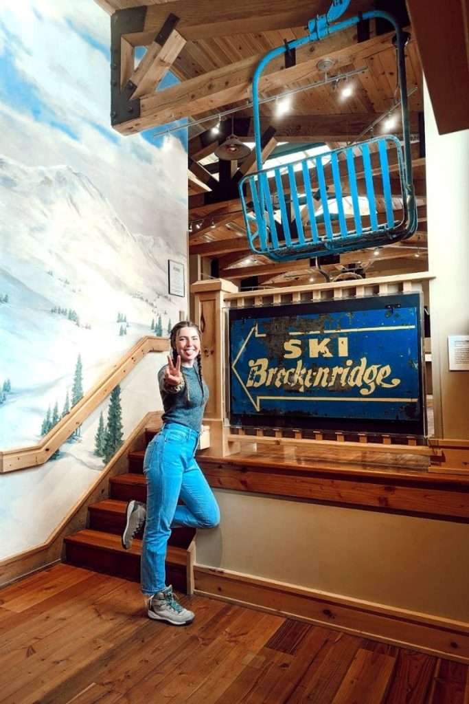 Visitor posing with a peace sign inside the Breckenridge Welcome Center in Colorado, standing beside a vintage blue “Ski Breckenridge” sign and ski lift chair display.