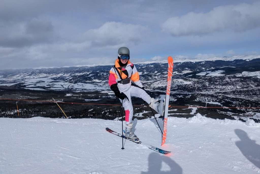 Skier posing playfully on one ski at the top of a snowy mountain in Breckenridge, Colorado, wearing a colorful retro ski suit with panoramic views of the Rockies in the background.