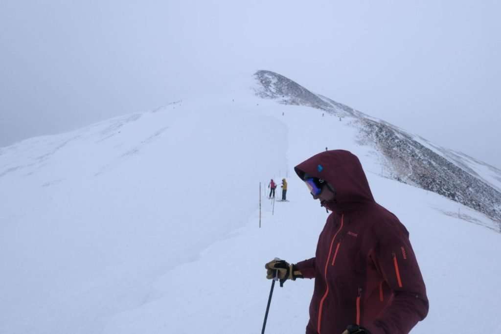 Skier wearing a red jacket and goggles standing near the top of a snowy ridge at Breckenridge Ski Resort in Colorado, with other skiers climbing toward the peak in the background.