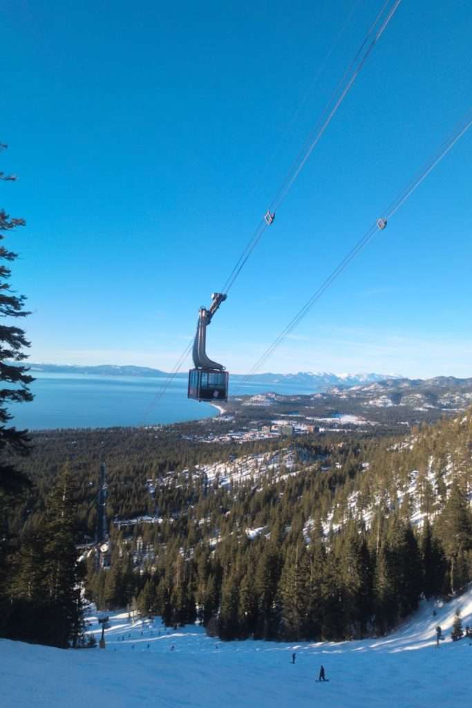 Gondola ascending over snow-covered trees and Lake Tahoe with clear blue skies at Heavenly Mountain Resort.