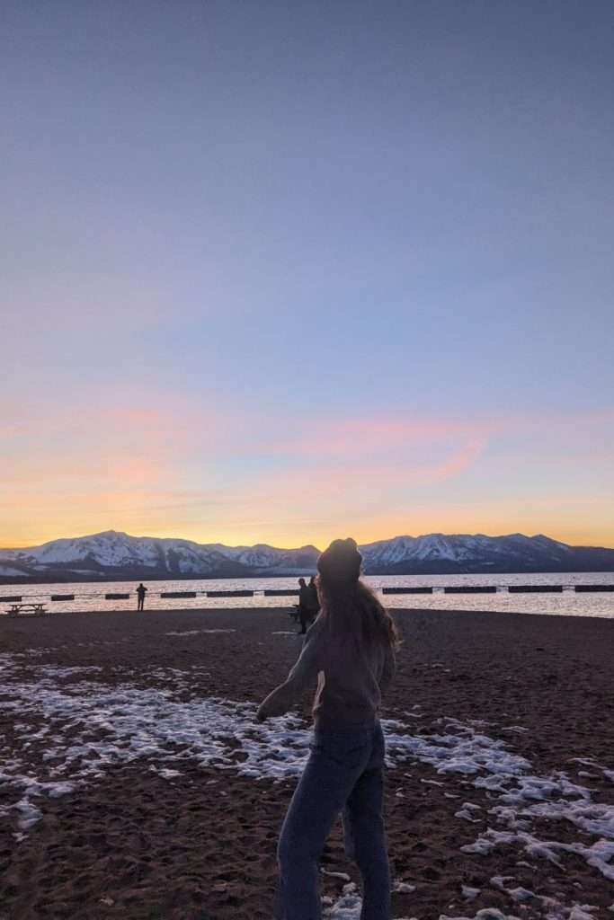 Silhouette of a woman standing on a snowy beach at sunset with mountain peaks and Lake Tahoe in the background.
