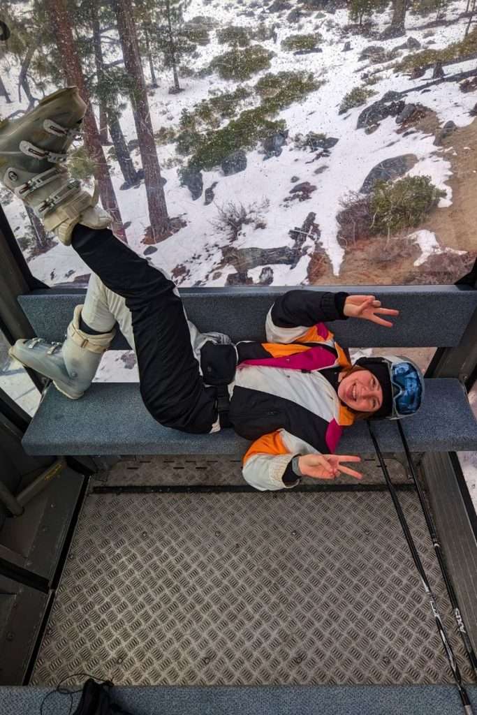 Traveler Liz lying playfully on a bench inside the Heavenly gondola with snowy trees visible below through the windows.
