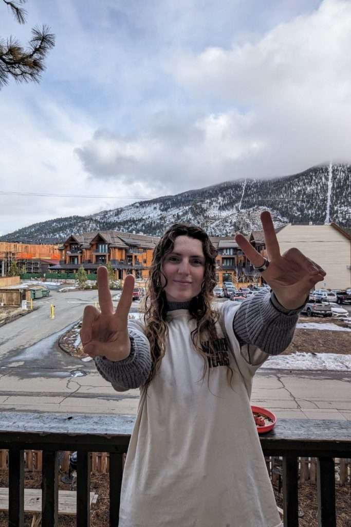 Traveler Liz flashing peace signs on the hostel balcony overlooking snowy mountains and winter lodges in South Lake Tahoe.
