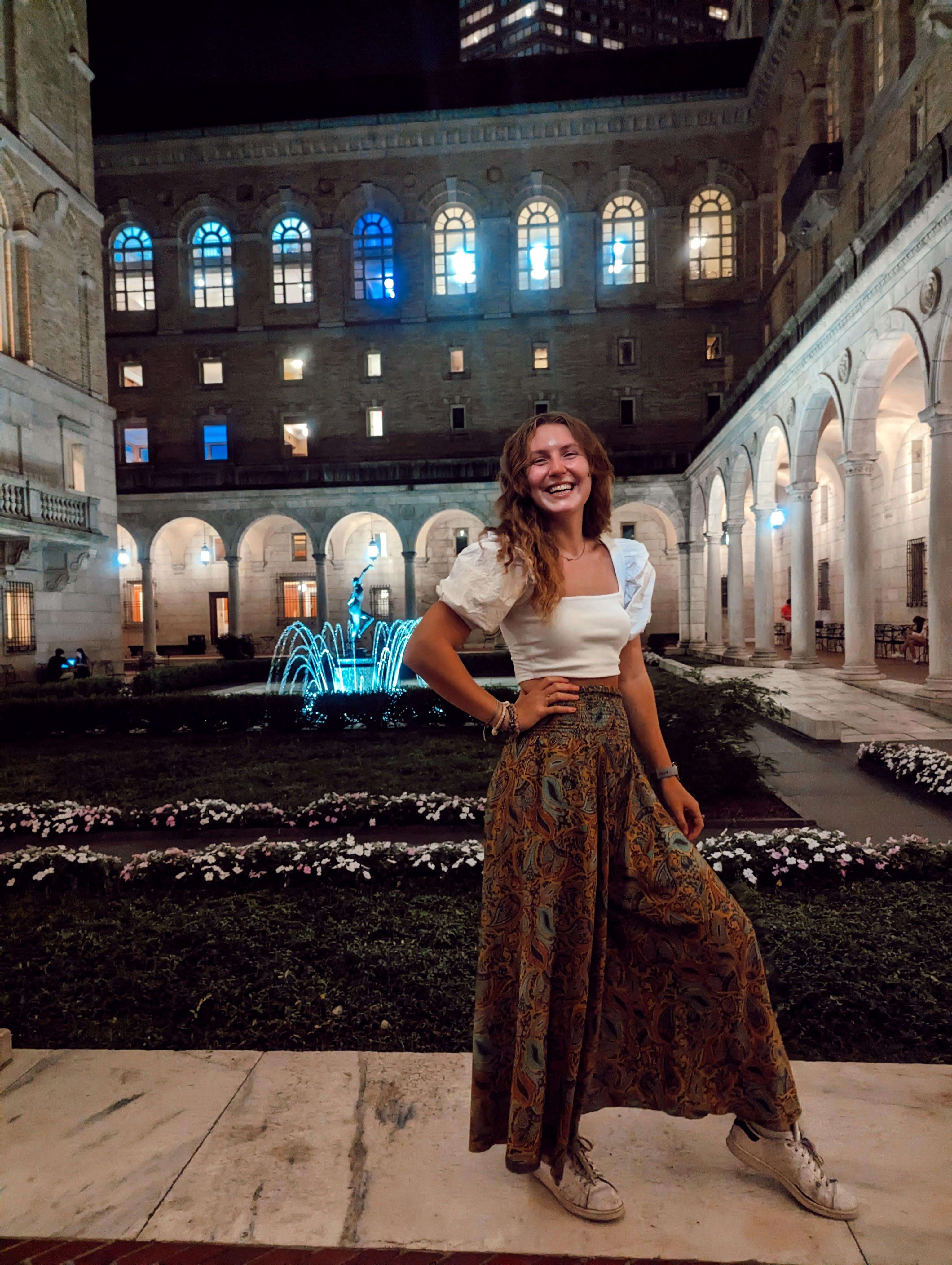 Me (Traveler Liz) standing in the Boston Public Library Courtyard at night with the fountain and many arches illuminated behind me.