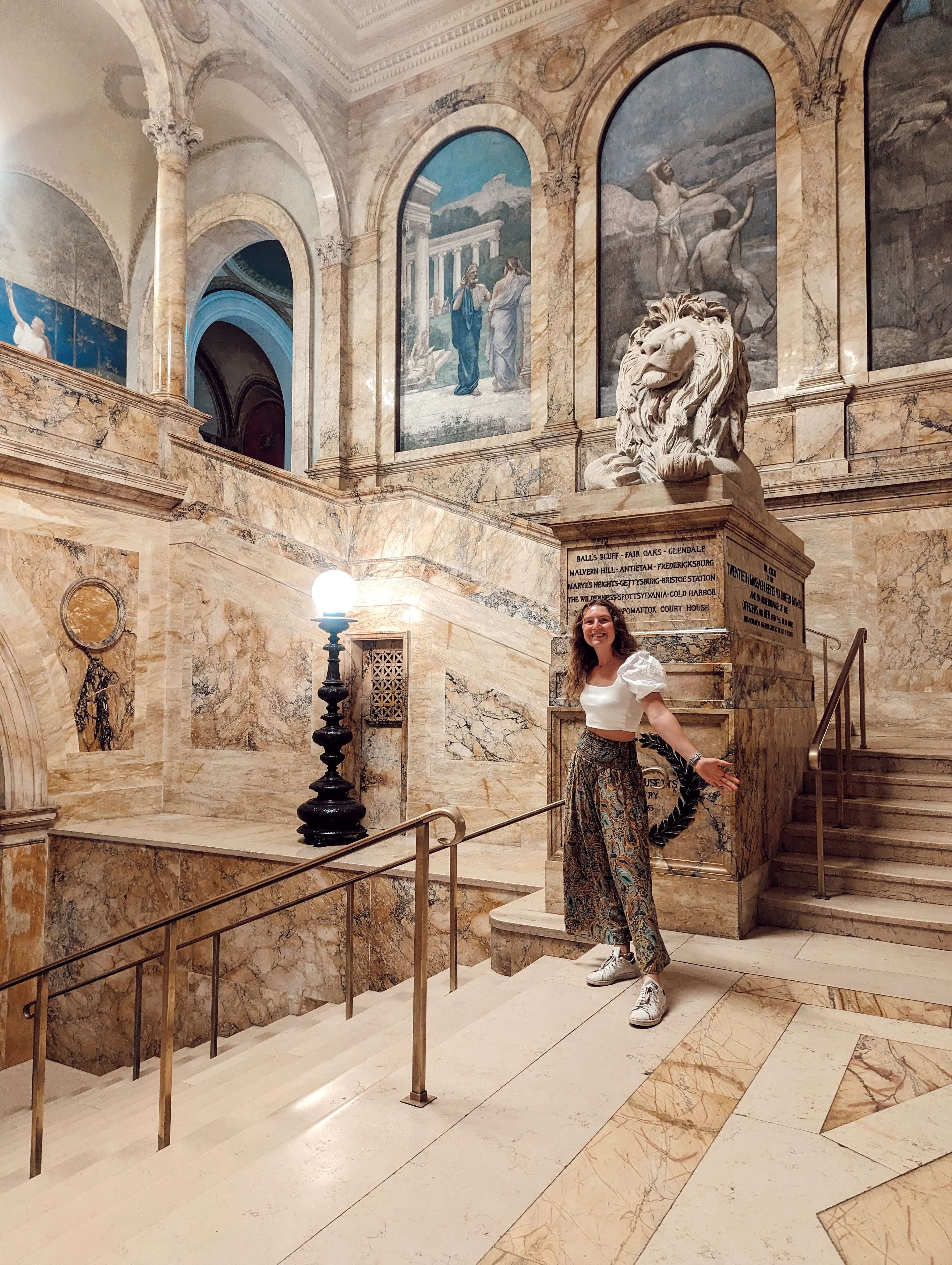 Me (Traveler Liz) standing in the stairs of the Boston Public Library, surrounded by the gorgeous marble and a lion statue.