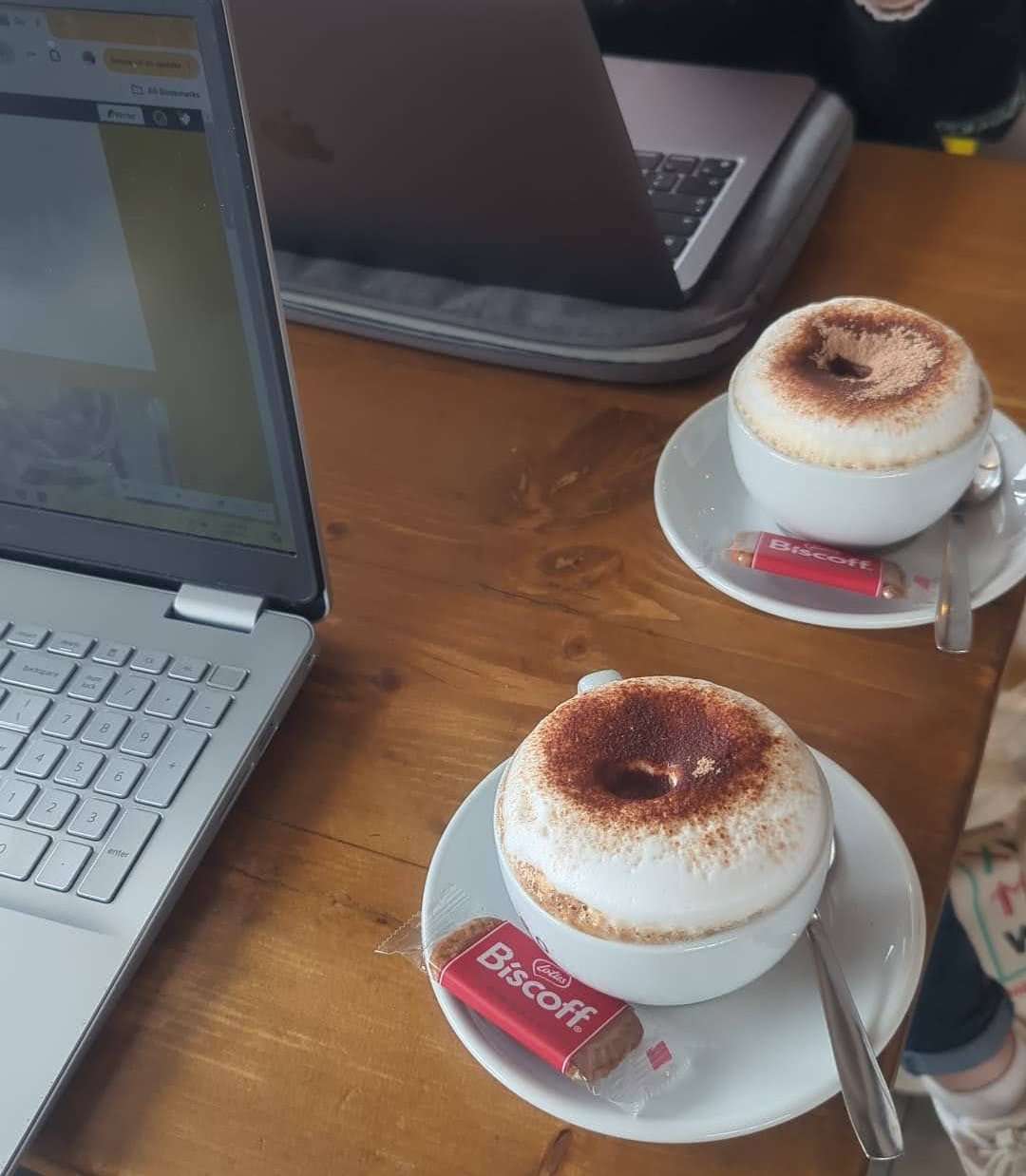 A couple of cappuccinos next to computers in a cafe.