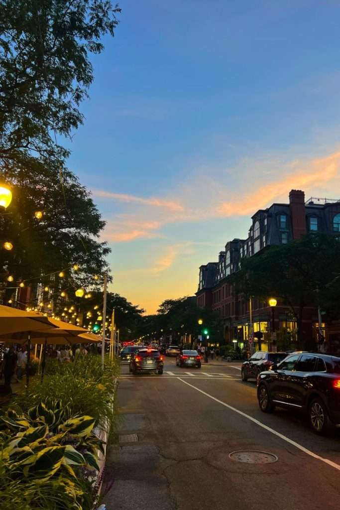 Boston Back Bay street at sunset with cars, string lights, and historic brownstone buildings glowing under a soft blue and pink sky.