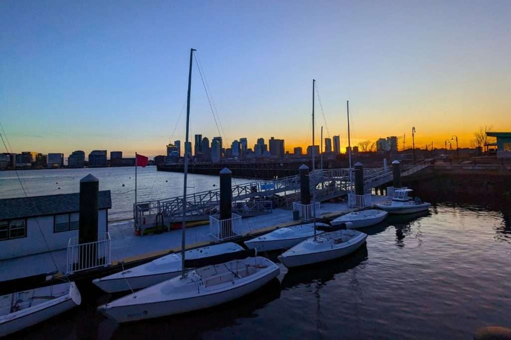 Sailboats docked along Boston Harbor at sunset with the city skyline glowing in the background.