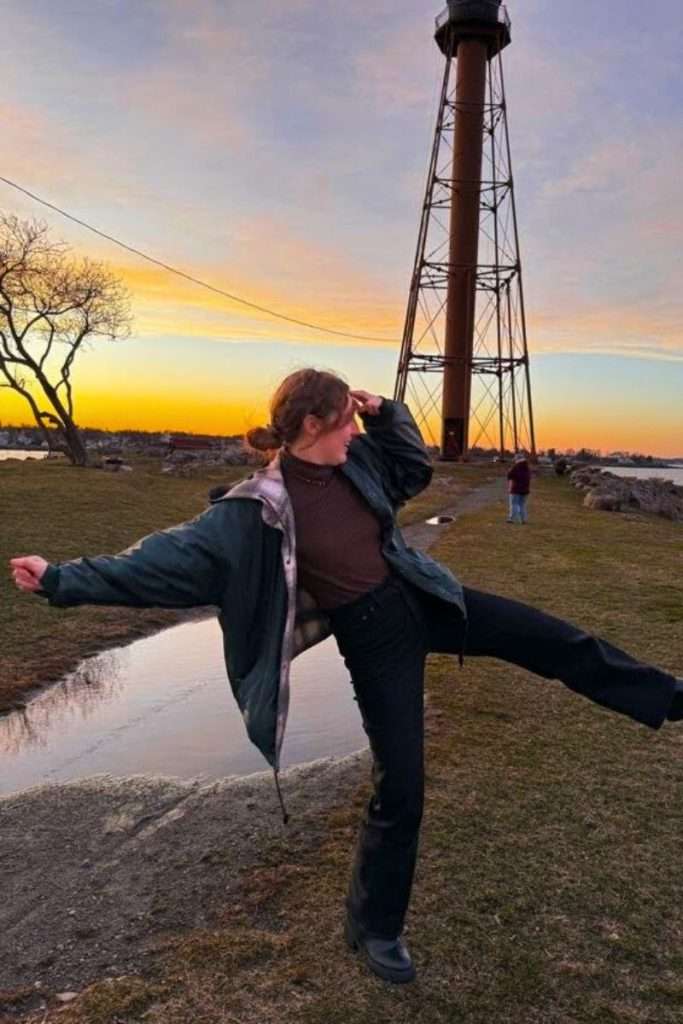 Traveler Liz playfully posing near a tall lighthouse during a colorful sunset in Boston.