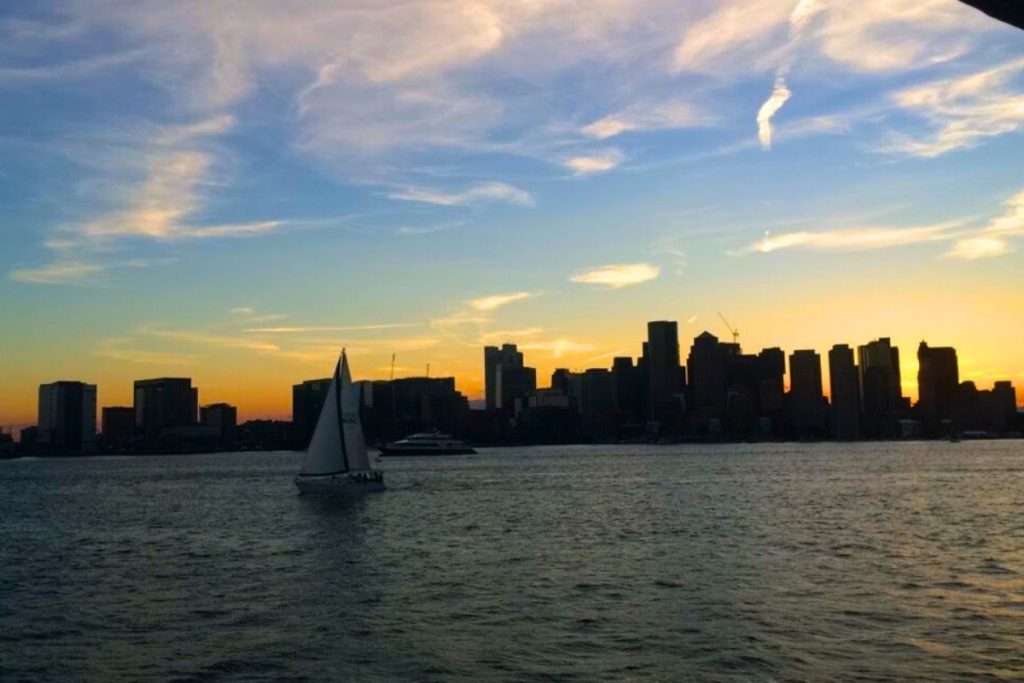 Sailboat gliding across Boston Harbor on a cruise at sunset with the city skyline silhouetted against a colorful sky.