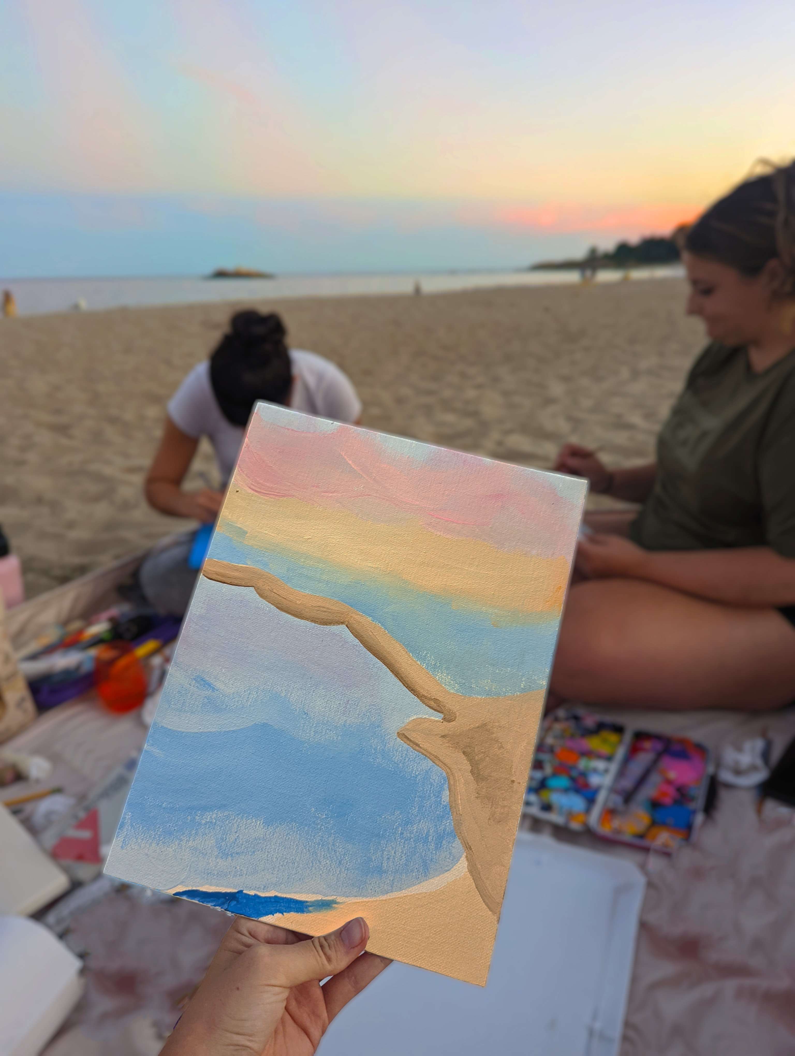 A painting of the beach with the sunset, in front of the sunset on that beach with light pink and blues.