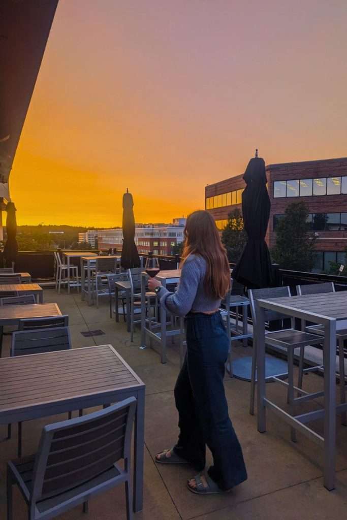 Traveler Liz holding a glass of wine on a rooftop terrace in Boston at sunset with orange sky in the background.