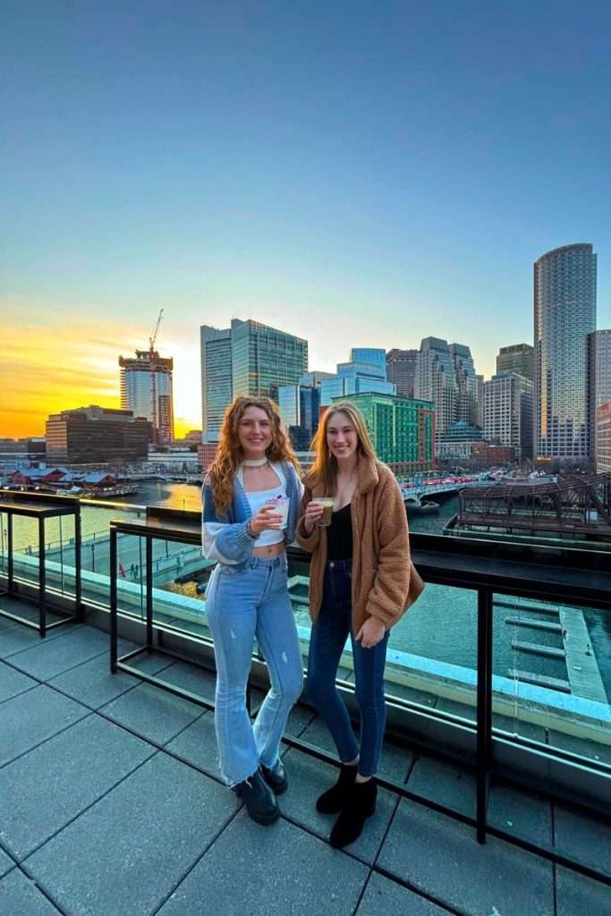 Two friends standing on a Boston rooftop at sunset enjoying cocktails with the orange sky and city skyline behind them.