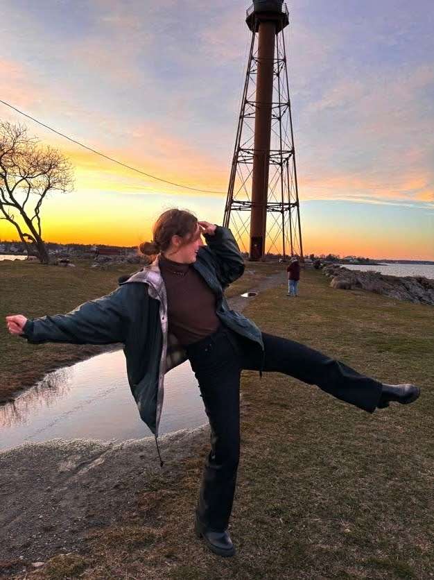 Me (Traveler Liz) with my leg up in front of a lighthouse with a ton of colors in the sky for the sunset in the back.