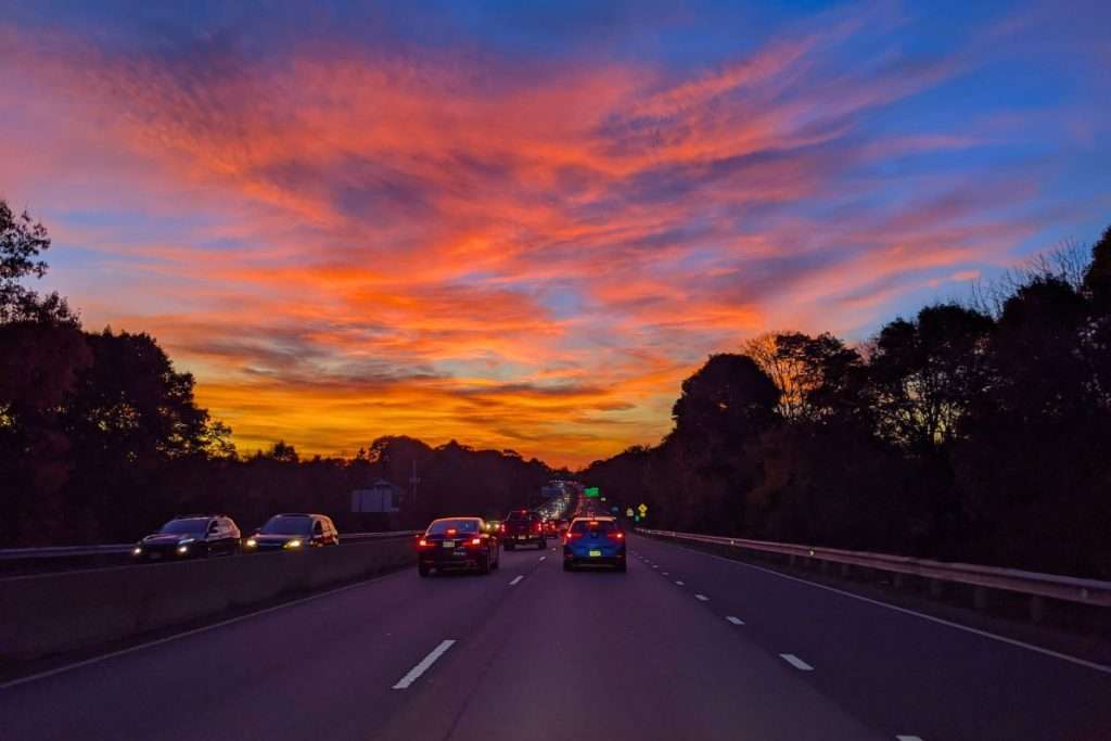 Cars driving west on a highway under a vibrant orange and purple Boston sunset sky.