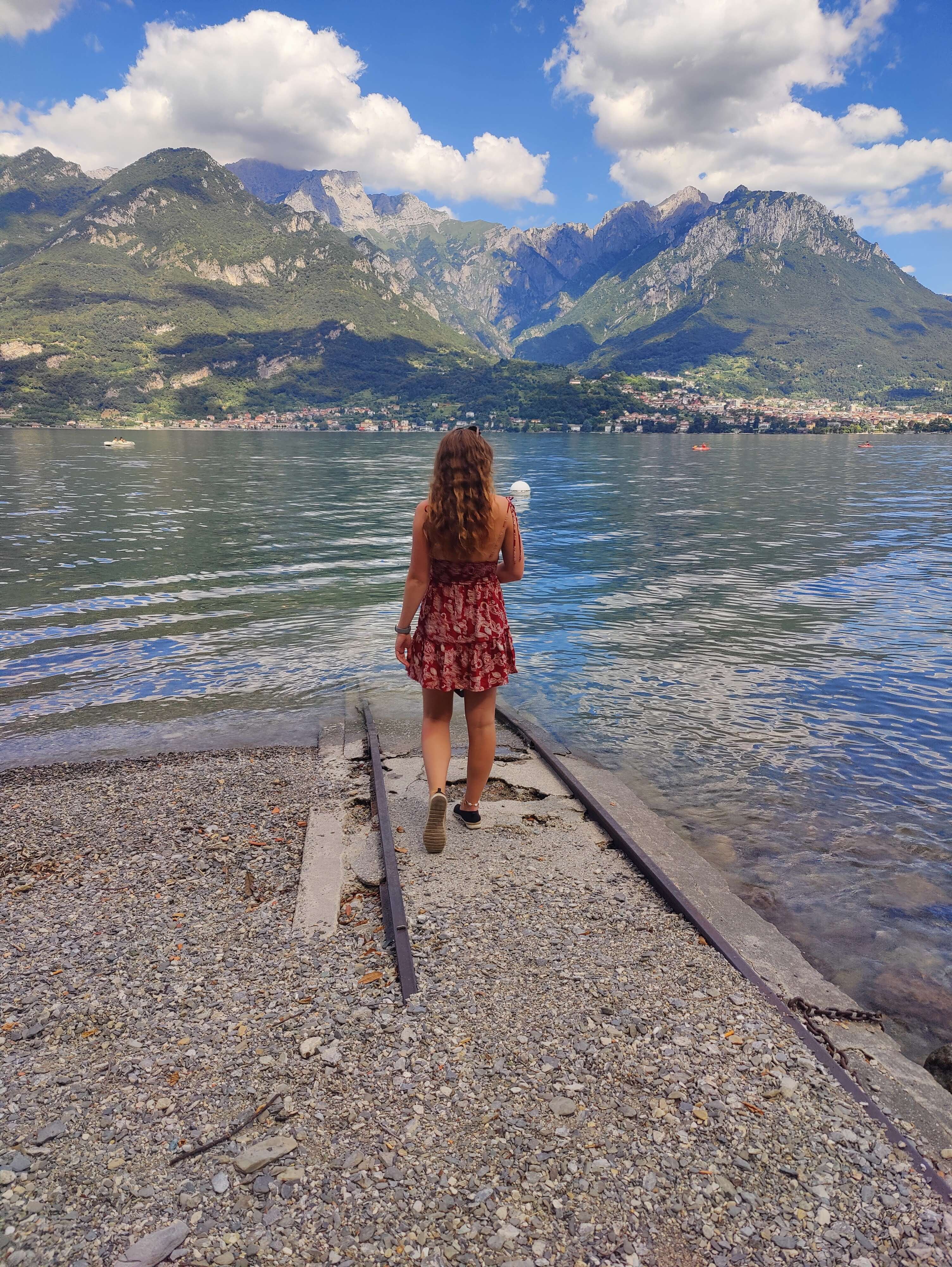A picture of me (Budget Traveler Liz), standing in front of Lake Como in Italy with a beautiful view of the mountains behind me and the reflection off the water.