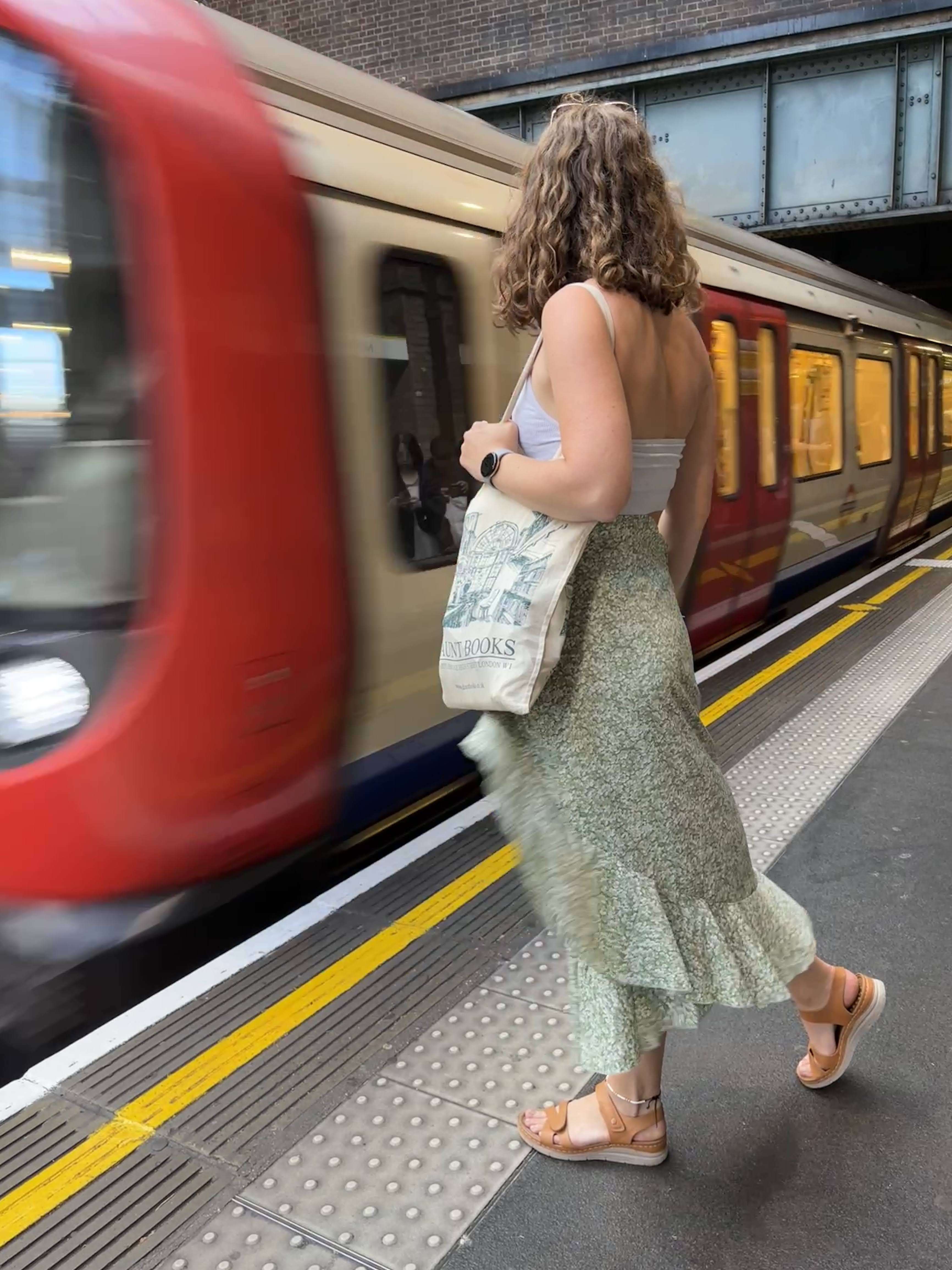 Me (Budget Traveler Liz) standing in front of the London tube as it pulls into the station with a flowy green skirt moving in the brief and my Daunt Books bag on my arm.