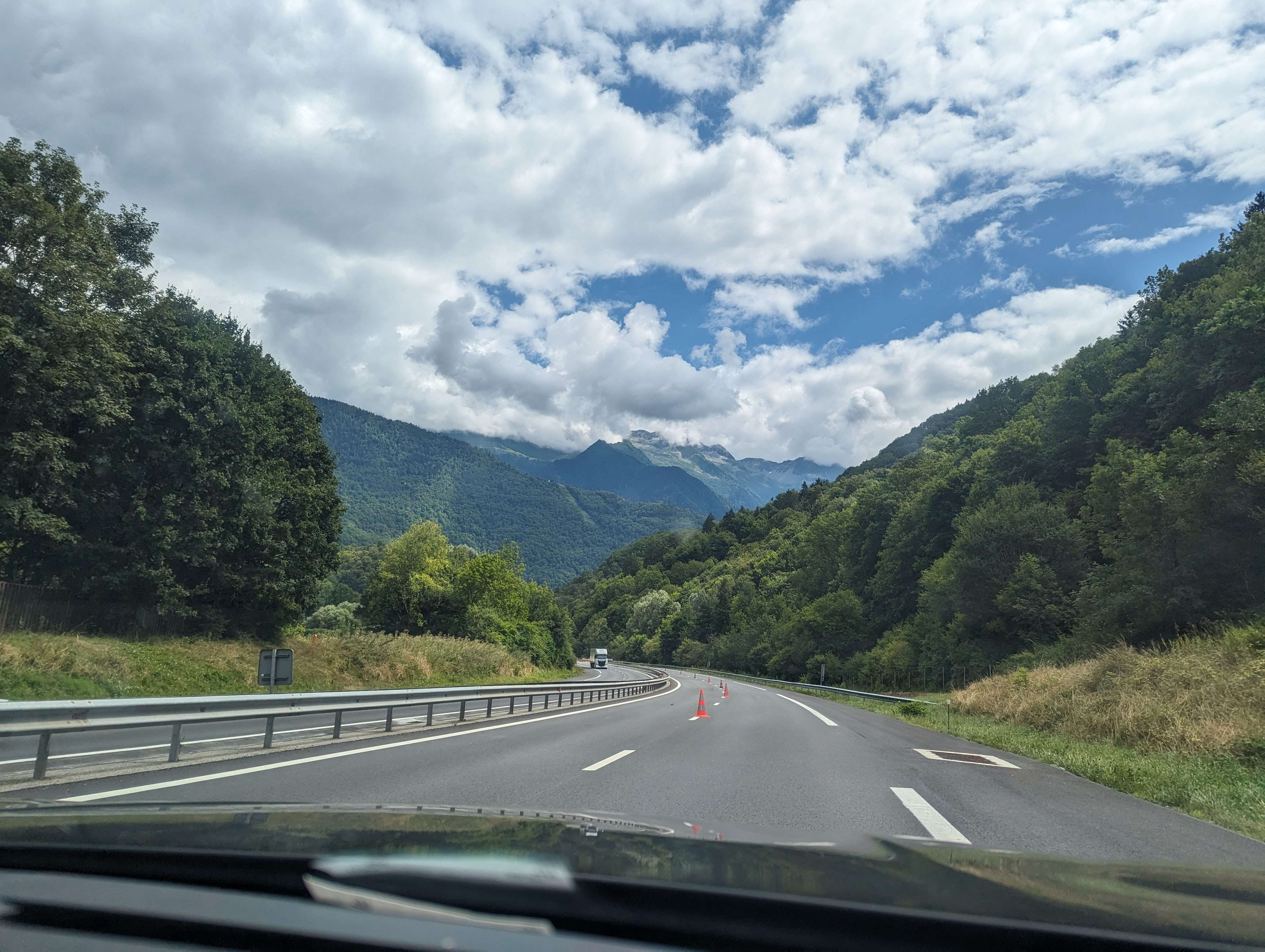 From the perspective of the car, driving along a road in France surrounded by trees and a mountain ahead in the distance.