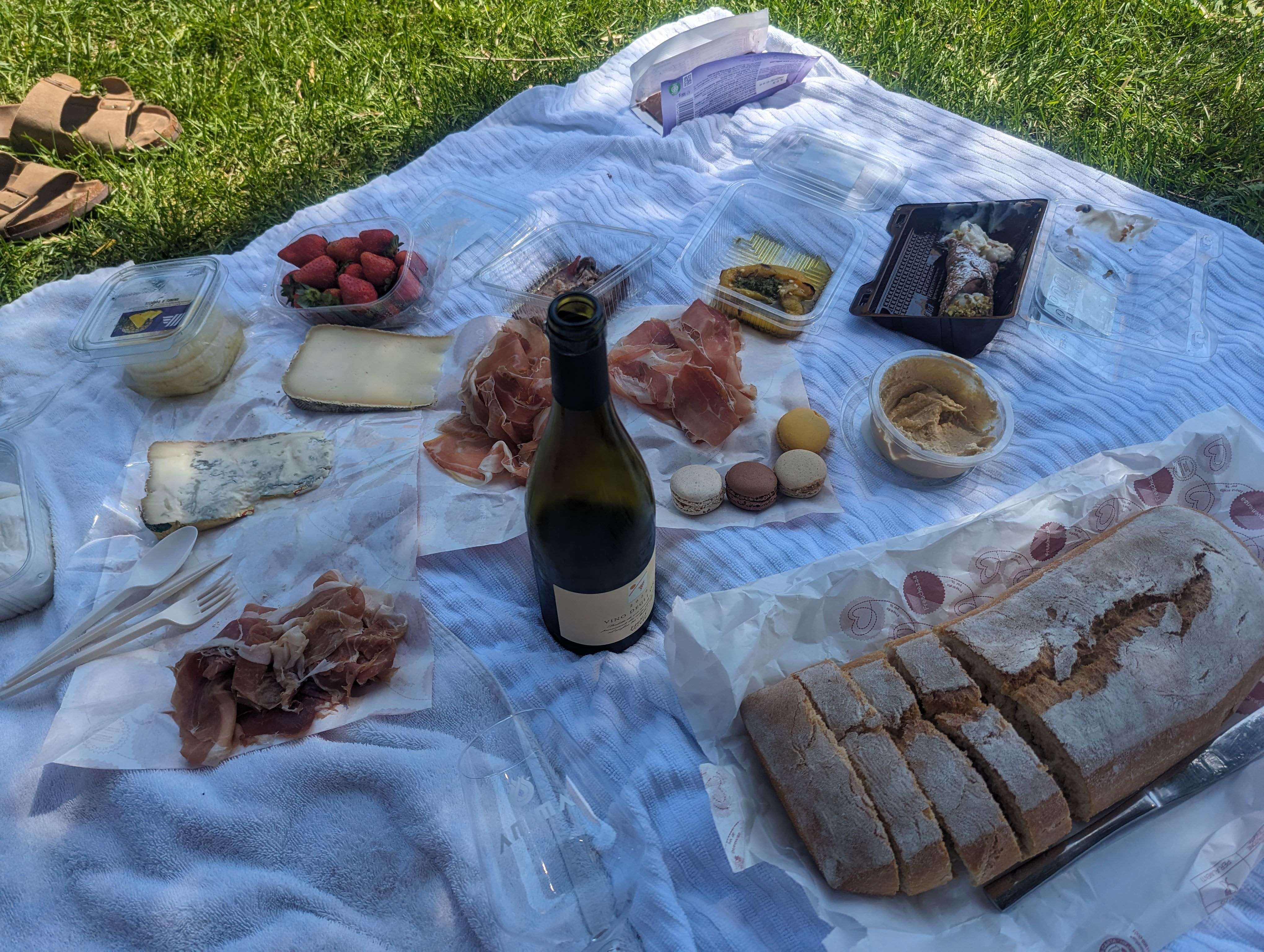 A picnic on the grass in France that includes bread, different types of cheese, a bottle of wine, strawberries and macaroons.
