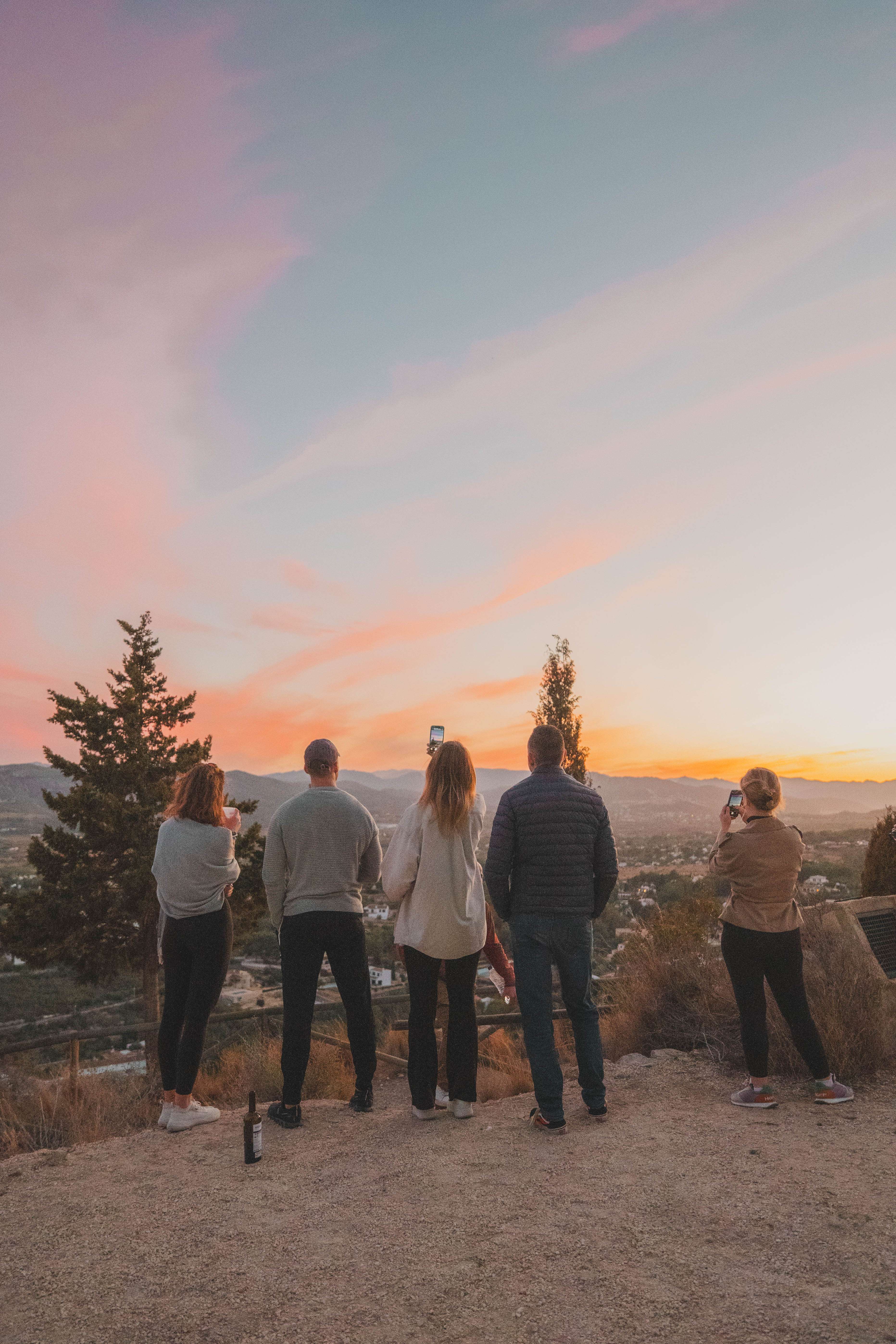 A few people standing around watching the sunset on top of a hill with some gorgeous pinks and oranges.