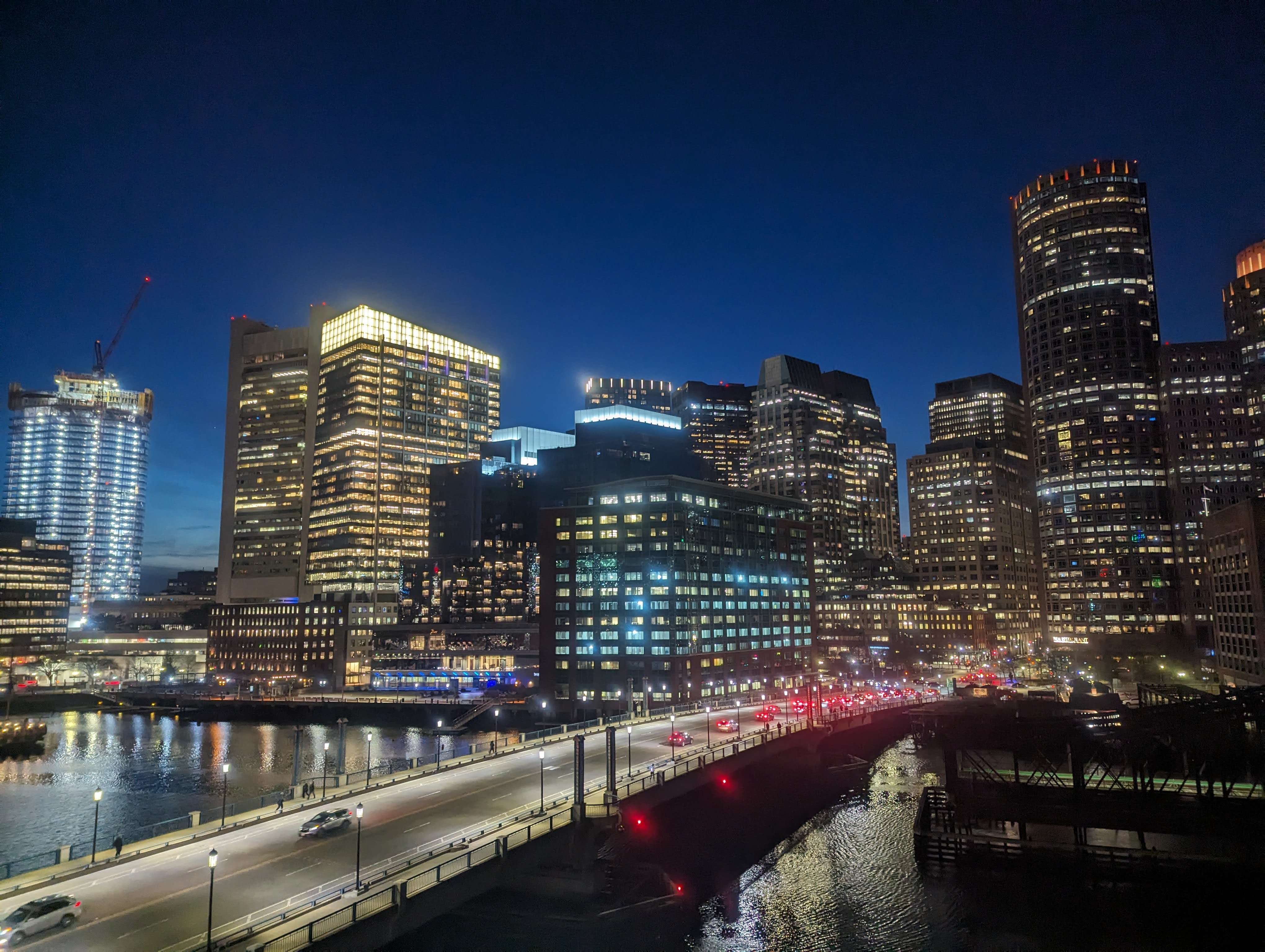 A view of Boston lit up at night with all the buildings and a illuminated road running through it.
