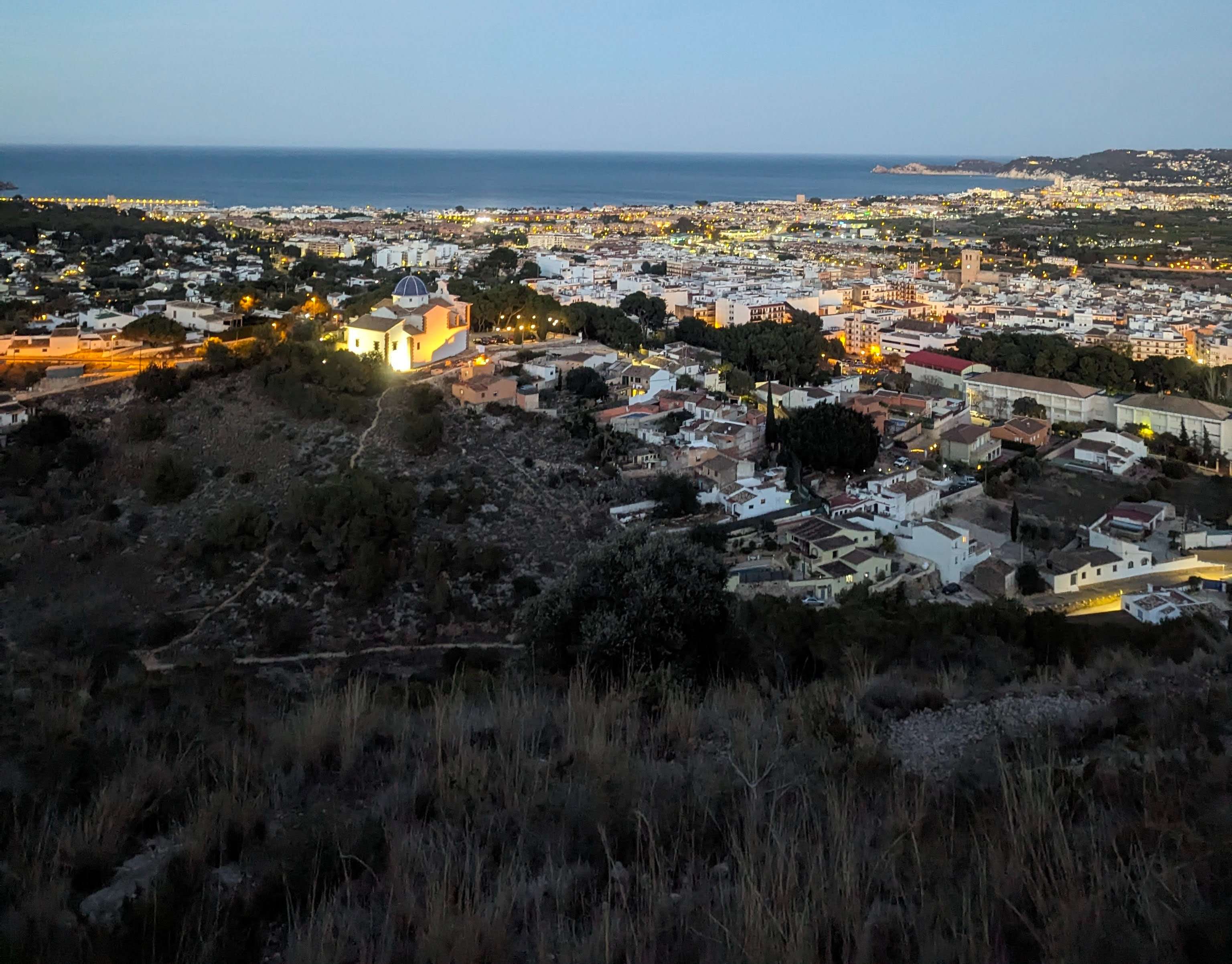 Jàvea/Xàbia at dusk from above with the city lit up with yellow and orange hues and the ocean in the background.