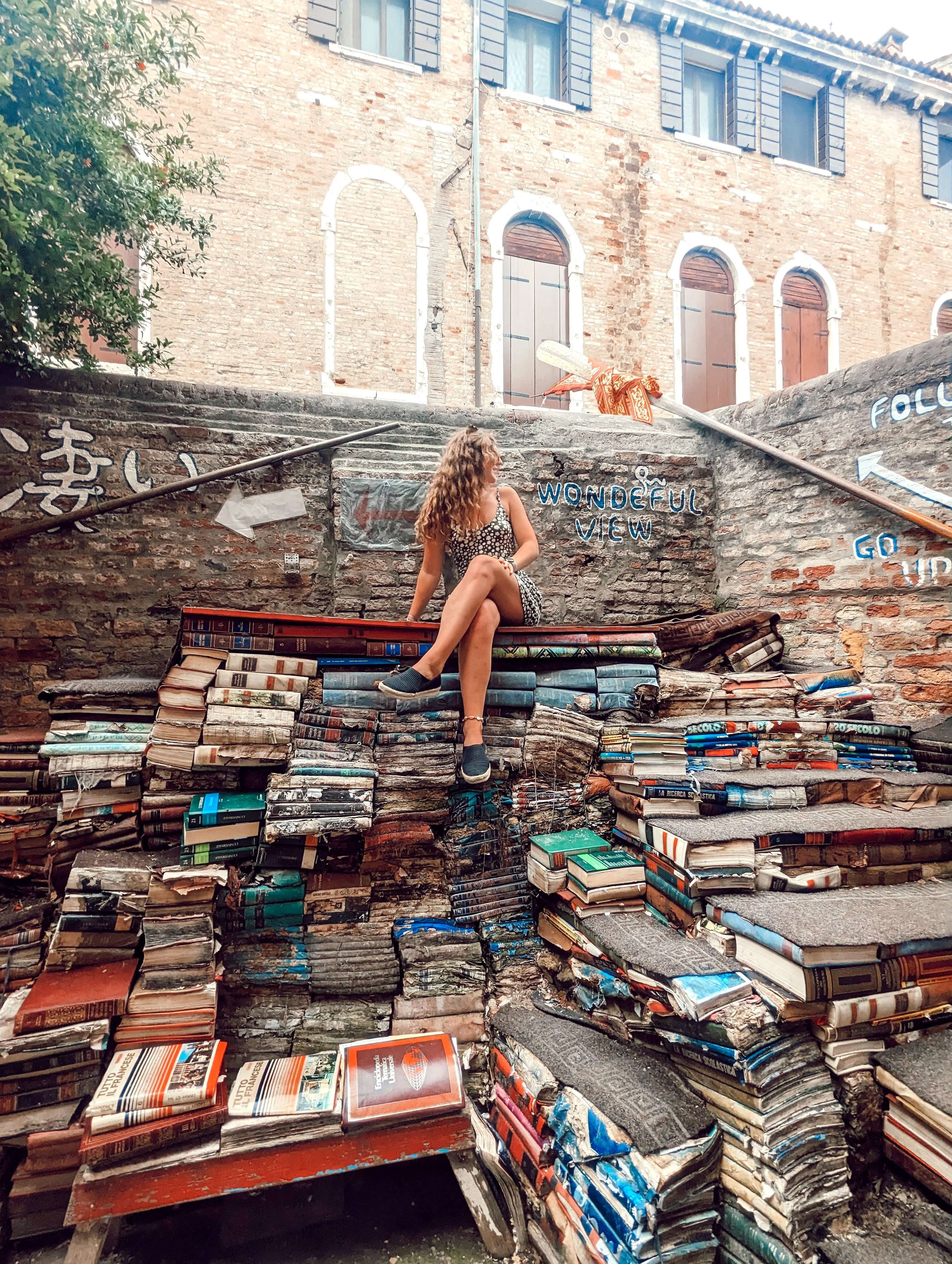 Me (Traveler Liz) sitting on a pile of books outside of Libreria Acqua Alta.