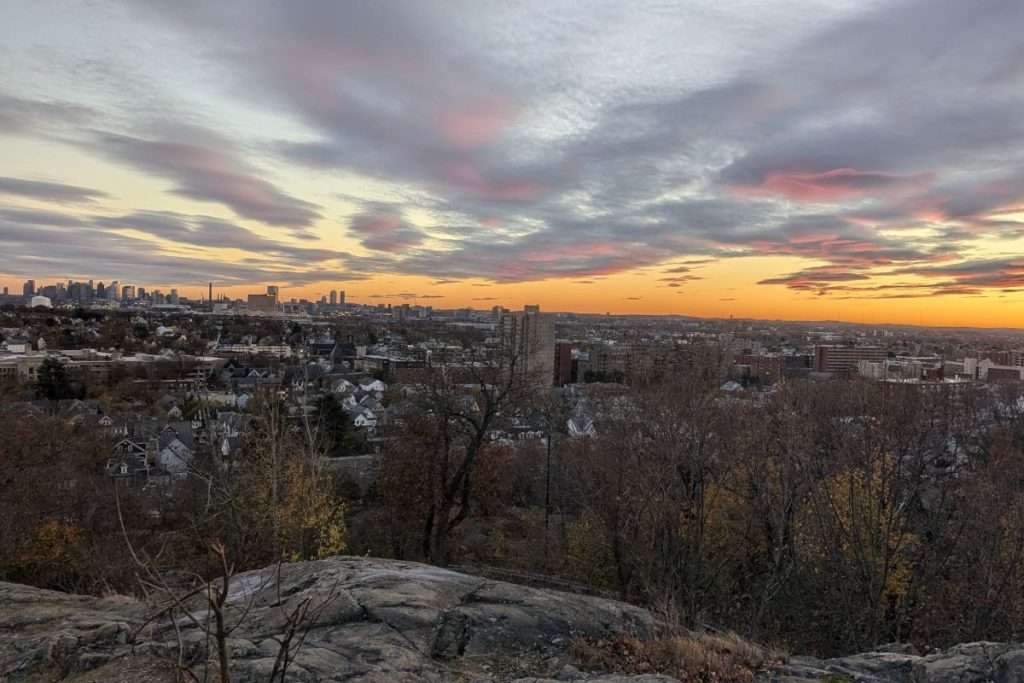 Sunset view over Boston skyline from a rocky overlook, with colorful clouds and city buildings in the distance