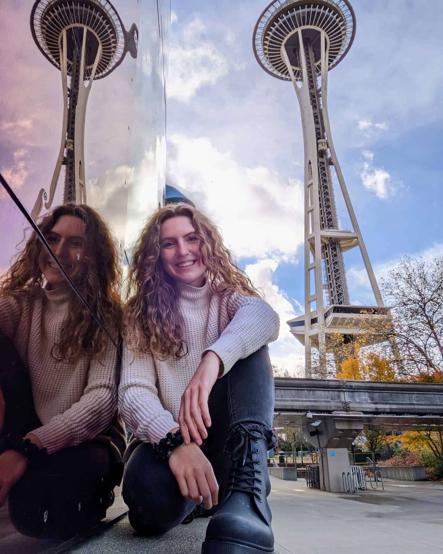 Me (Traveler Liz) kneeling next to the Museum of Pop Culture with the Space Needle behind me.