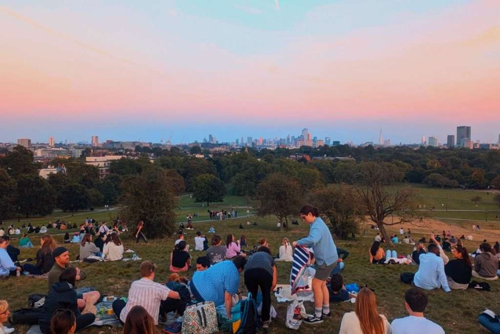 Crowds gathered on Primrose Hill in London at sunset, enjoying a picnic with views of the city skyline under a pink and blue sky.