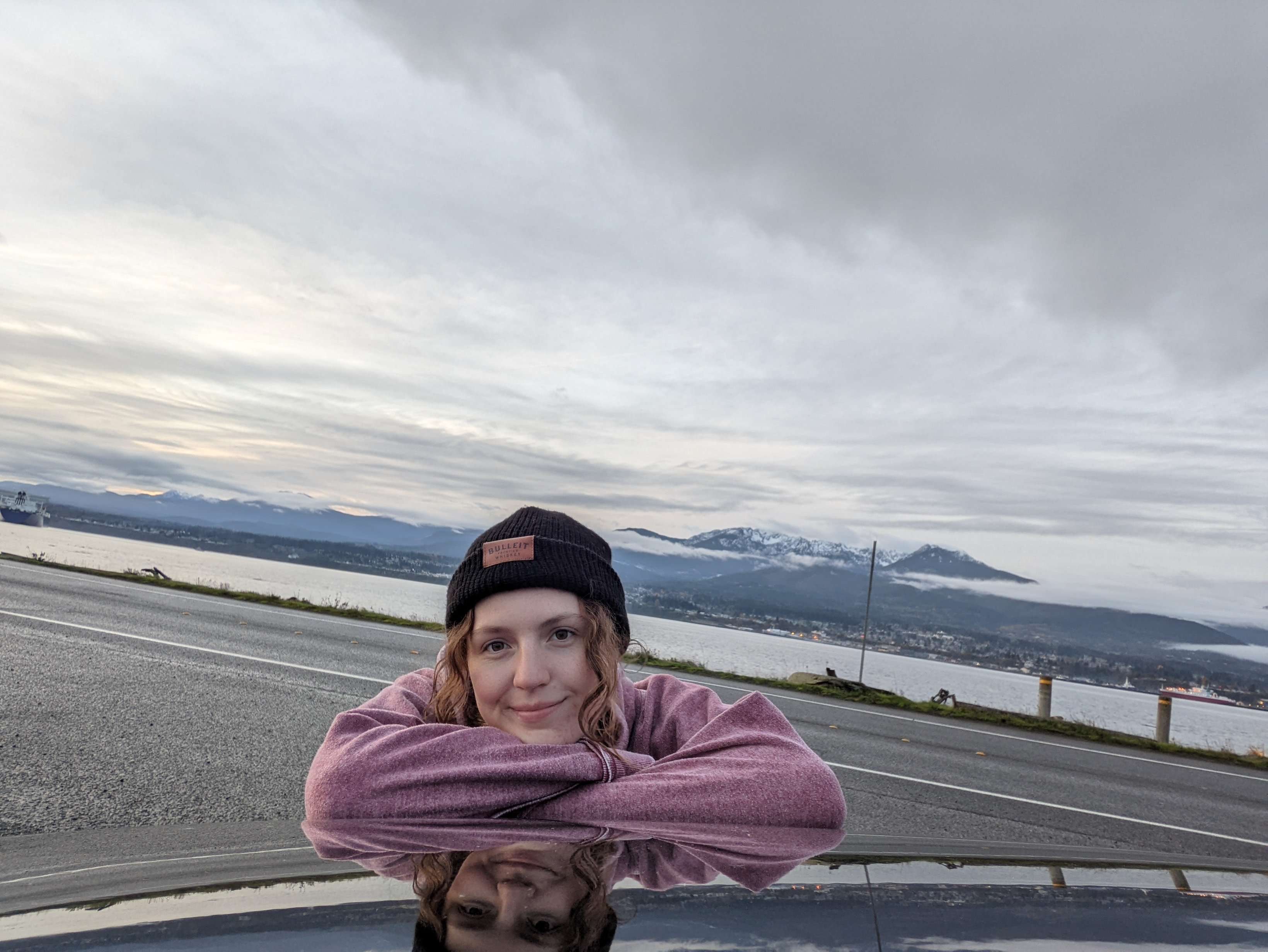Me leaning on my arms on my car with the mountains behind me and my reflection on the hood.