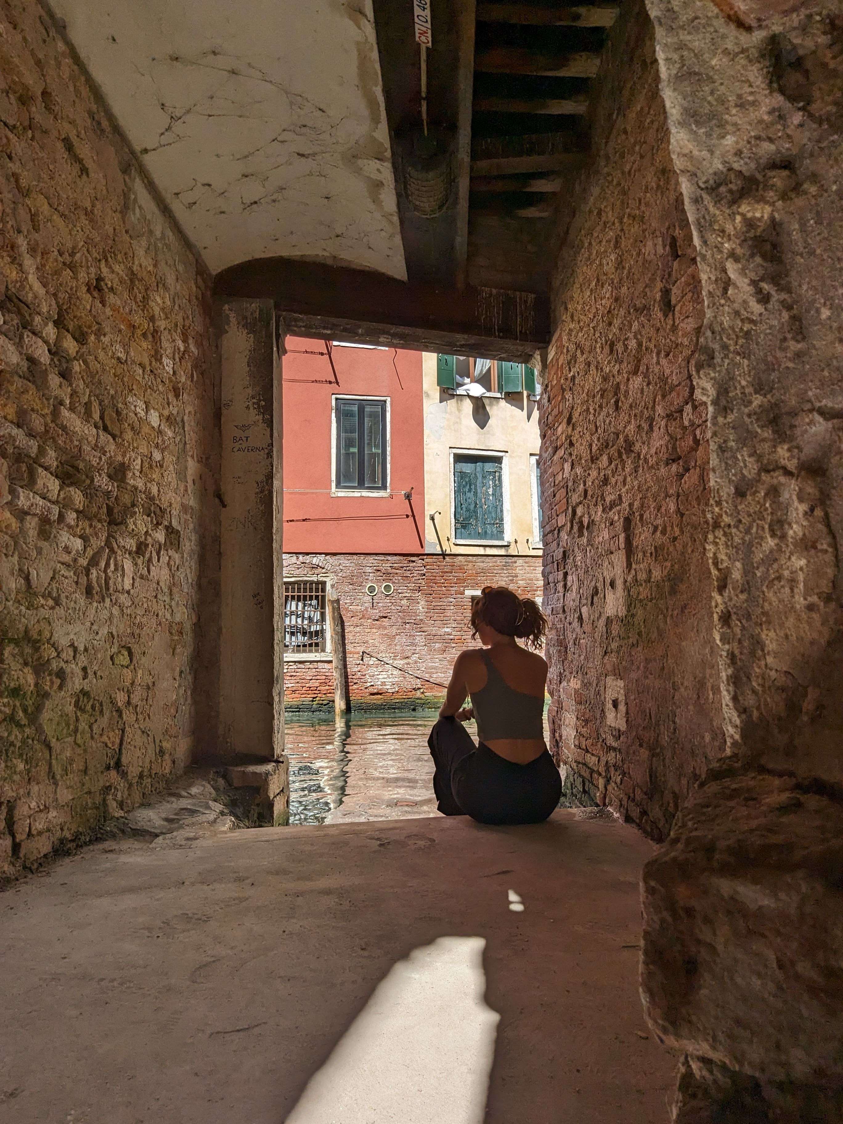 Me (Traveler Liz) sitting at the end of the narrowest street in Venice overlooking a canal.