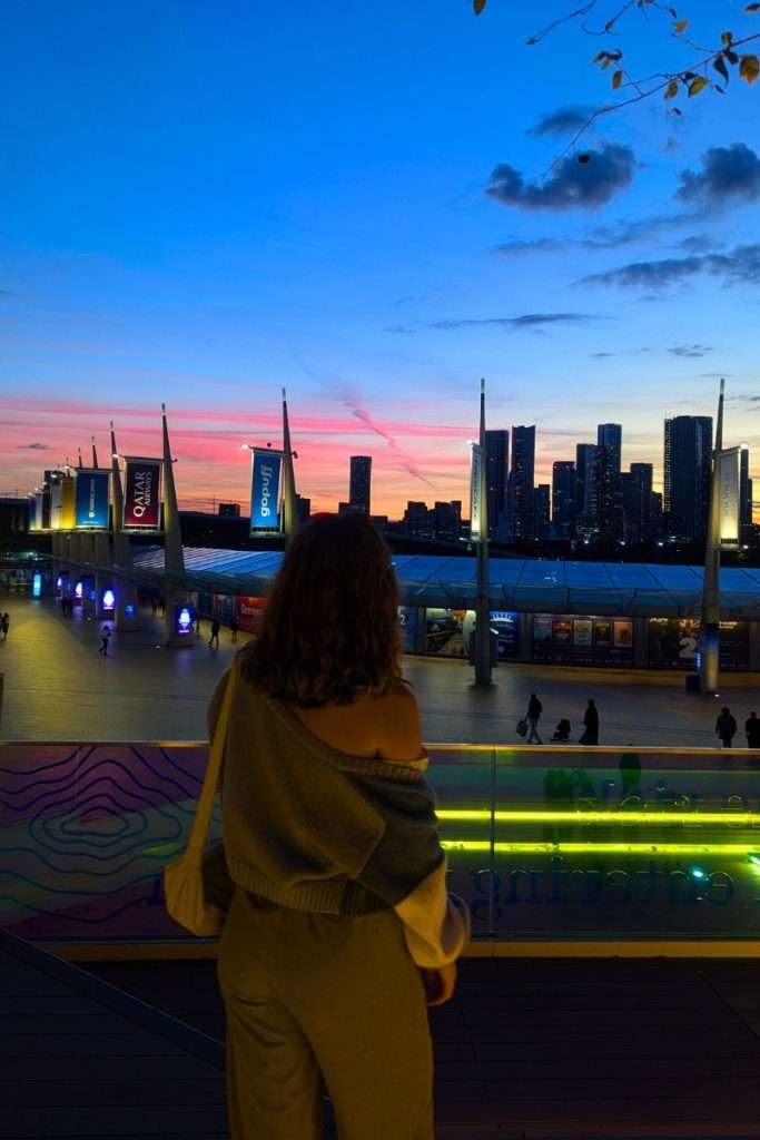 Person watching a colorful sunset over London’s skyline from The Tide walkway near The O2, with city lights beginning to glow.