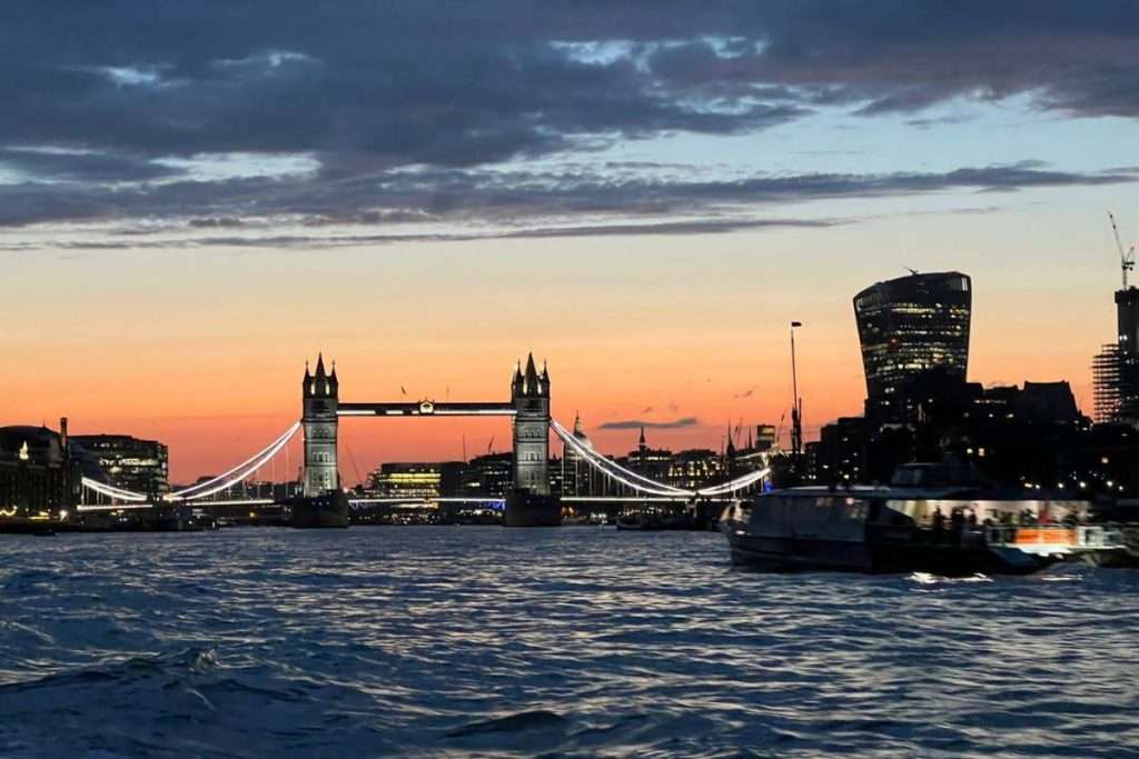 Tower Bridge in London at sunset with lights reflecting on the River Thames and a boat passing in the foreground.