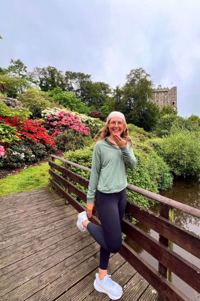 Liz standing outside Blarney Castle on a bridge surrounded by flowers and greenery in Ireland.