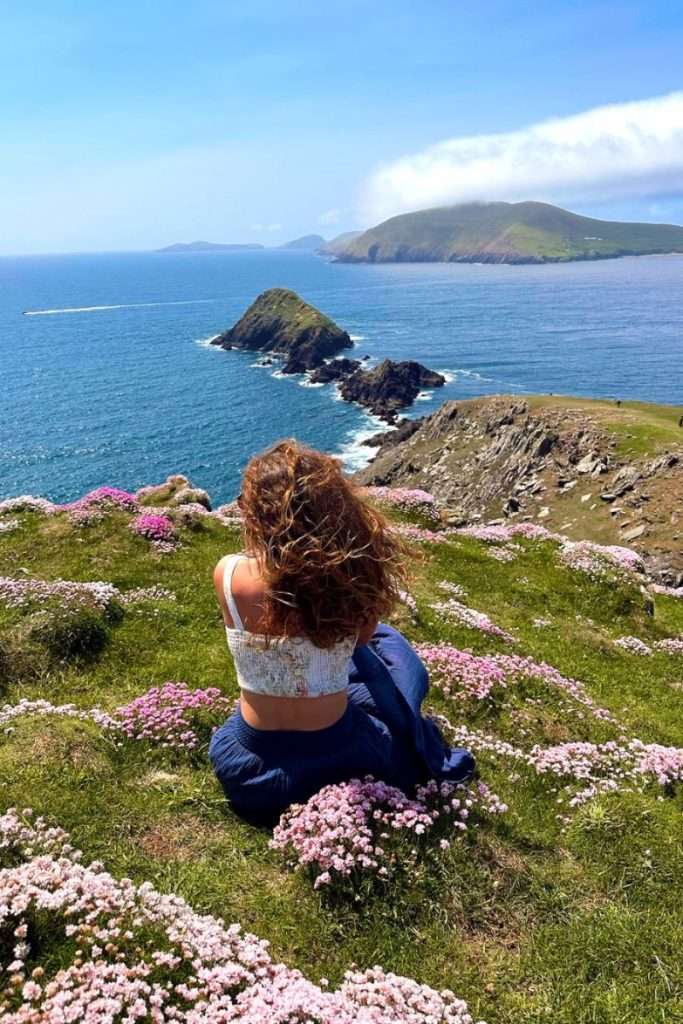 Liz sitting in a field in Ireland near Dingle surrounded by flowers and staring at the ocean.