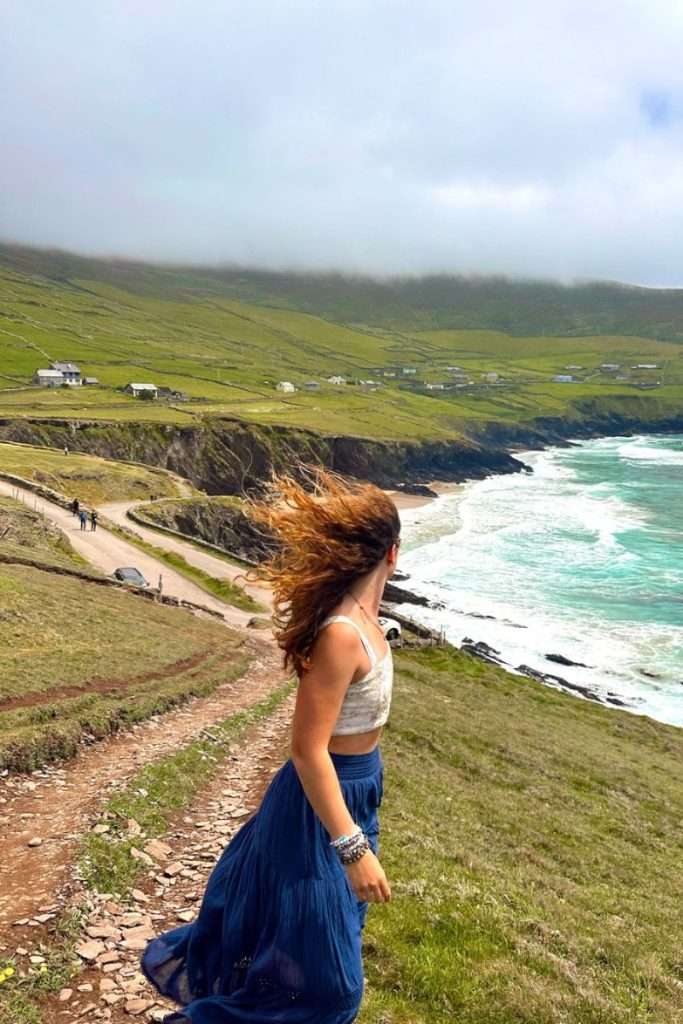 Liz standing on a windy cliffside in Ireland during her roadtrip looking down on a beautiful blue beach.