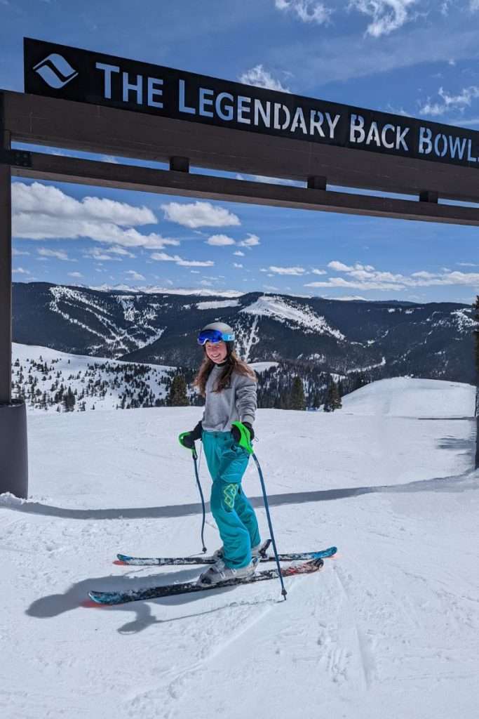 Skier smiling under ‘The Legendary Back Bowls’ sign at Vail Resort, Colorado, surrounded by snowy mountain peaks, one of the best Epic Pass resorts in Colorado.