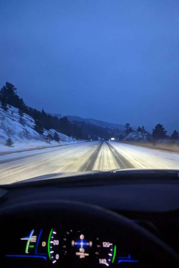 Driving on a snowy Colorado mountain road at dawn with headlights illuminating the icy highway, a reminder to prepare for winter conditions before a Colorado ski trip.