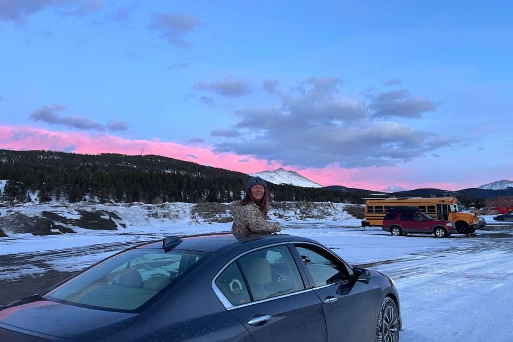 Traveler smiling from the sunroof of a rental car at sunset in the Colorado mountains, with pink skies and snow-covered peaks in the background in the free parking lot in Breckenridge.