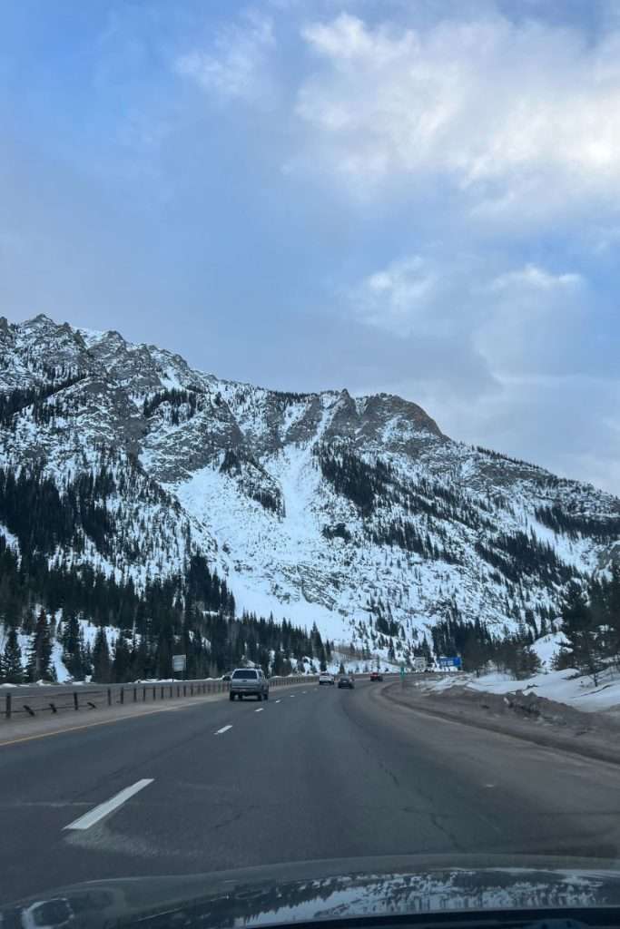 Cars driving along I-70 through the Colorado Rockies, with snow-covered mountains and pine trees lining the highway, a scenic winter drive between ski resorts.