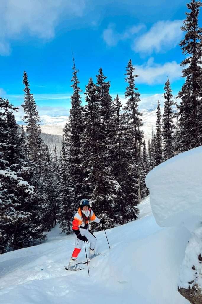 Skier making their way through fresh powder and pine trees on a bluebird day in Colorado, enjoying backcountry-style terrain on a ski trip.