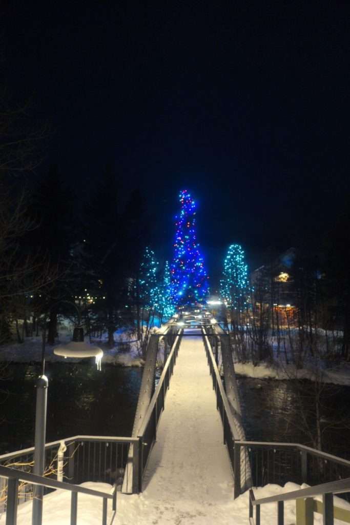 Snowy footbridge leading to brightly lit blue Christmas trees behind The Pad hotel in Silverthorne, Colorado, a festive winter night scene in Summit County.