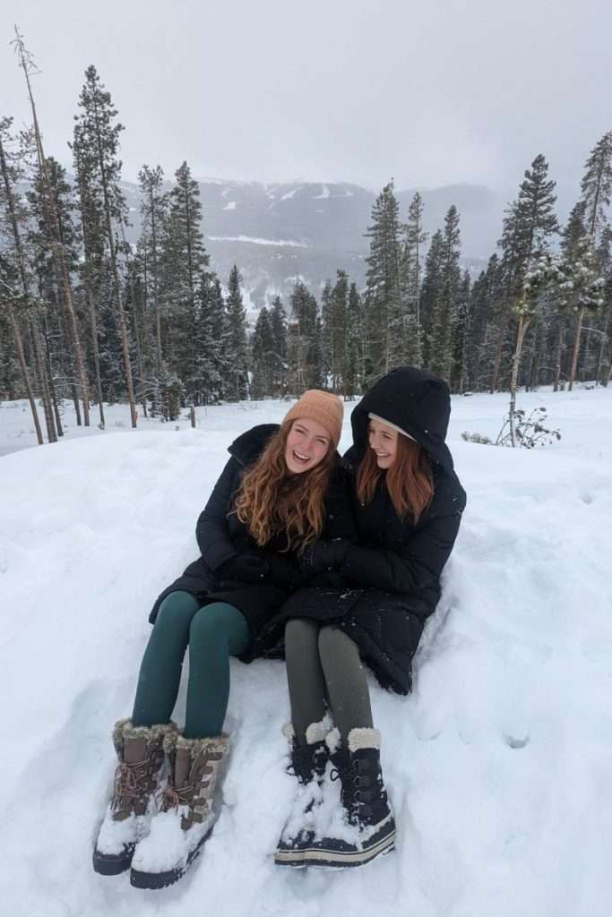Two sisters sitting in the snow in Colorado wearing winter coats, leggings, and snow boots, a cozy outfit example for what to pack on a Colorado ski trip.