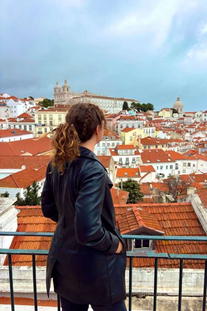 Traveler Liz with curly brown hair in a black jacket looking out over the red rooftops and historic buildings of Lisbon from a high viewpoint on a cloudy day.