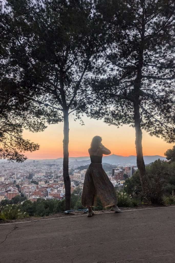 Traveler Liz standing between two trees overlooking Barcelona at sunset from Mirador del Nen de la Rutlla, capturing the panoramic city view.