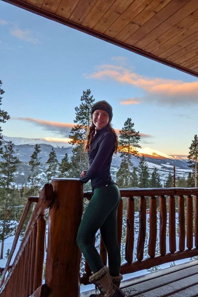 Woman standing on a snowy cabin balcony in Colorado at sunrise, wearing a mock neck long sleeve, winter boots, and a warm base layer bottom, a cozy outfit example for a ski trip packing list.