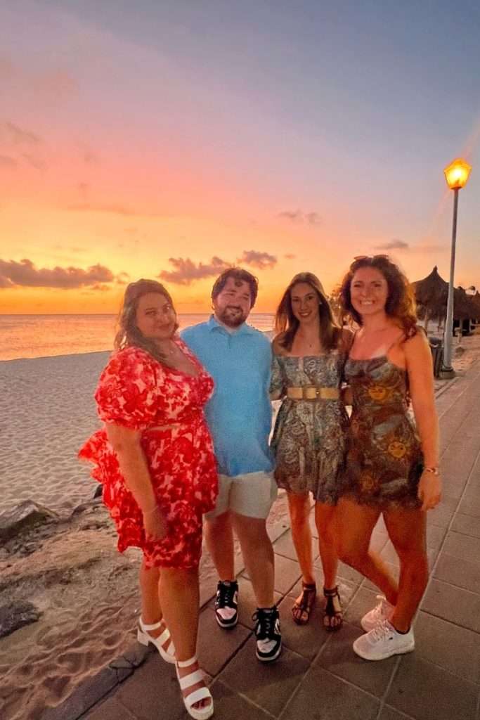 Group of friends standing together on a beachside walkway in Aruba at sunset, with colorful sky, sand, and lamplights in the background.