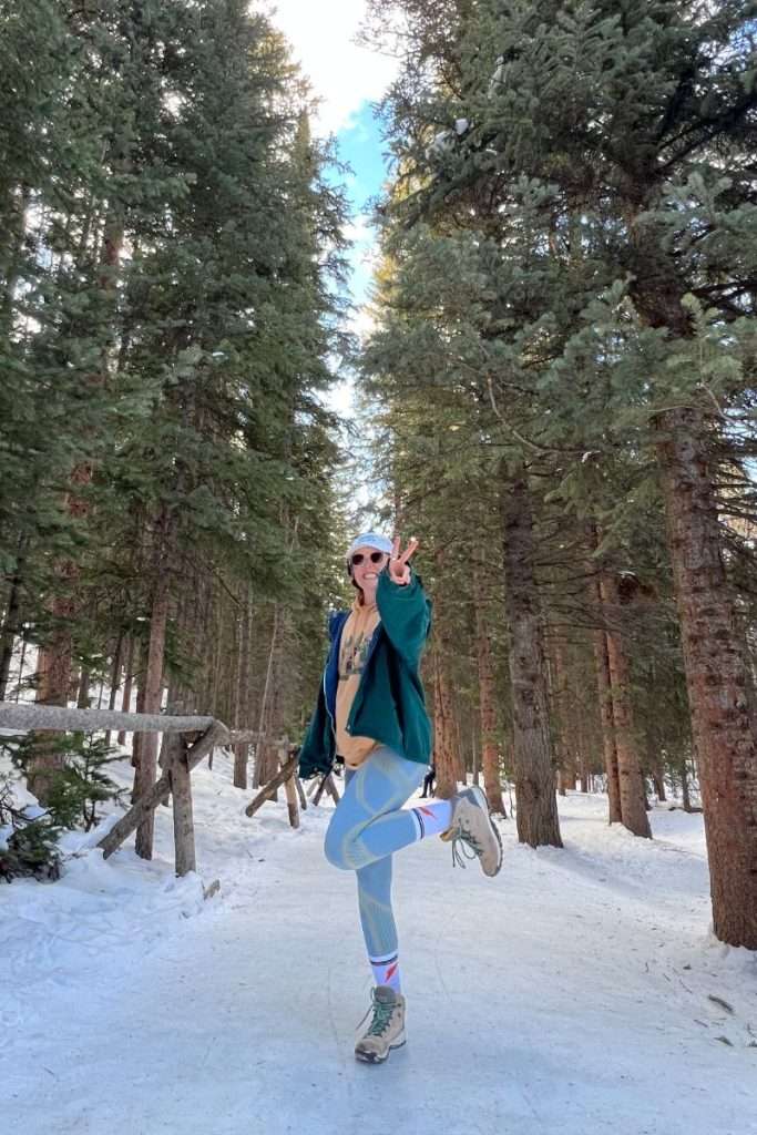 Woman posing on a snowy trail in Colorado wearing hiking boots, leggings, and layered winter clothes, showing practical footwear for a ski trip or winter hike.