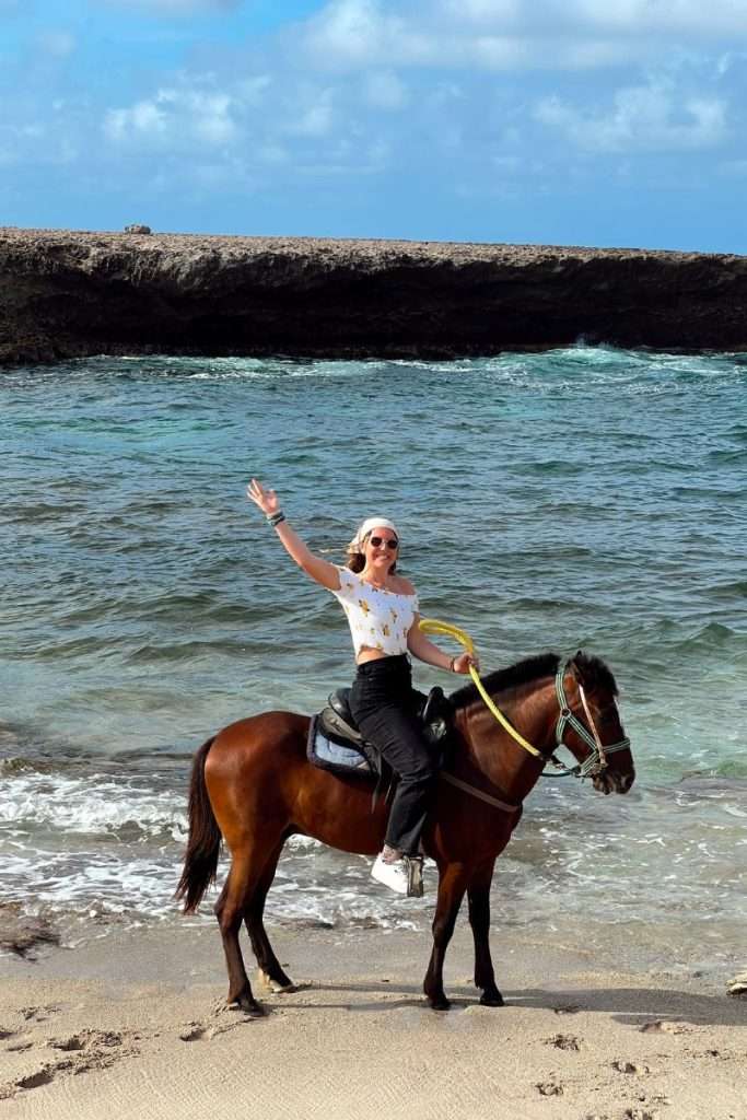 Person horseback riding along the shoreline in Aruba, smiling and waving with ocean waves and rocky coastline in the background.